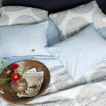 A John Robshaw Kama Light Indigo Organic Sheet Set bed with pillows and a magazine on it. - 28362869604398