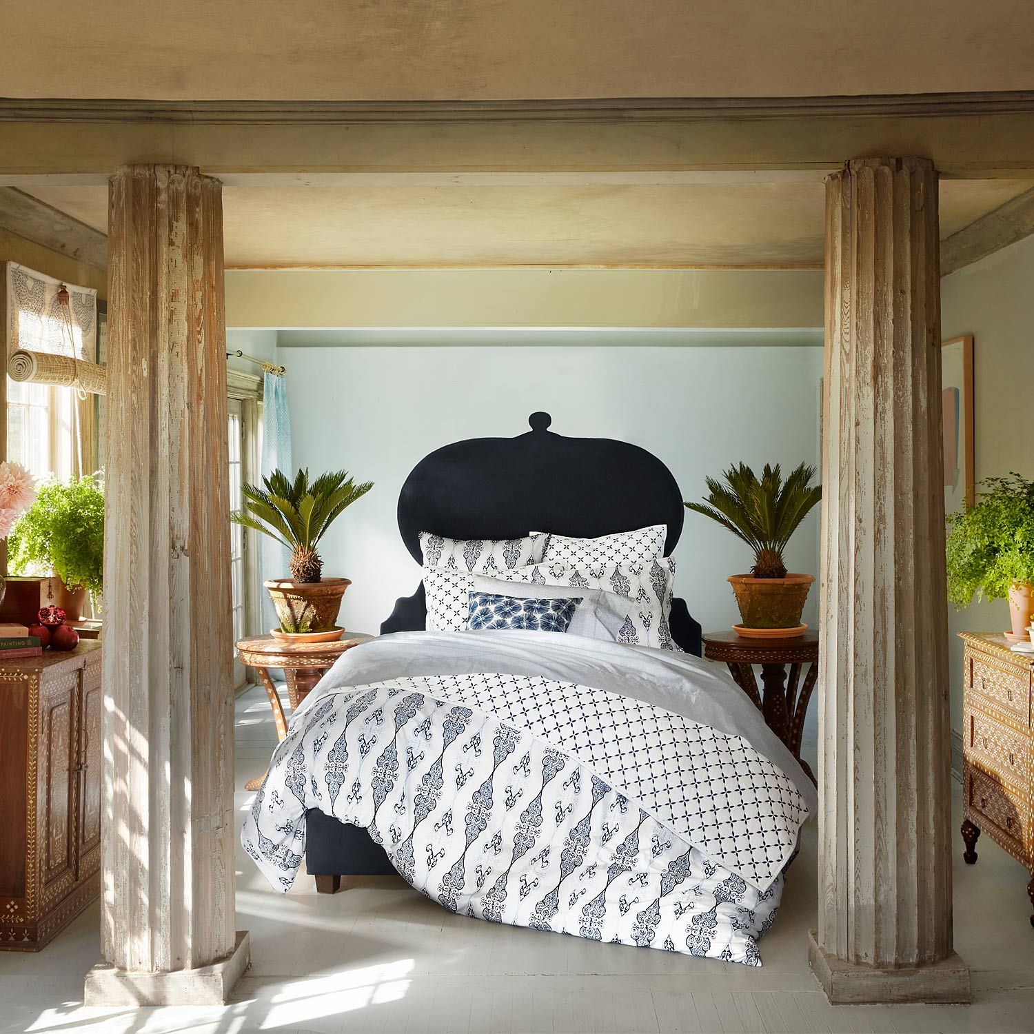 A bedroom with a Layla Indigo Quilt by John Robshaw dressed in soft cotton voile sheets, accompanied by a stylish bedside table.